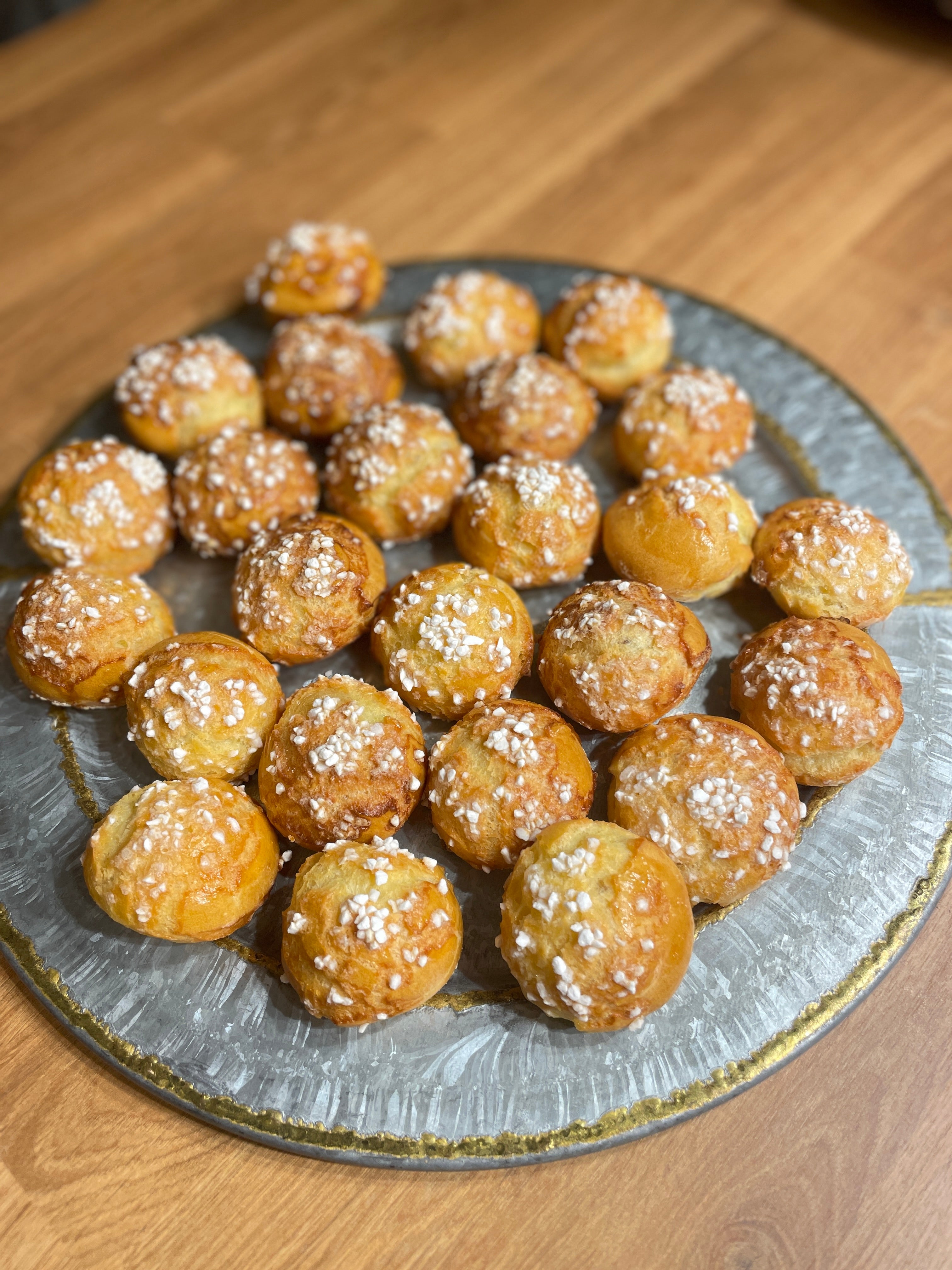 A round silver serving tray filled with golden brown chouquettes, topped with pearl sugar, placed on a wooden table
