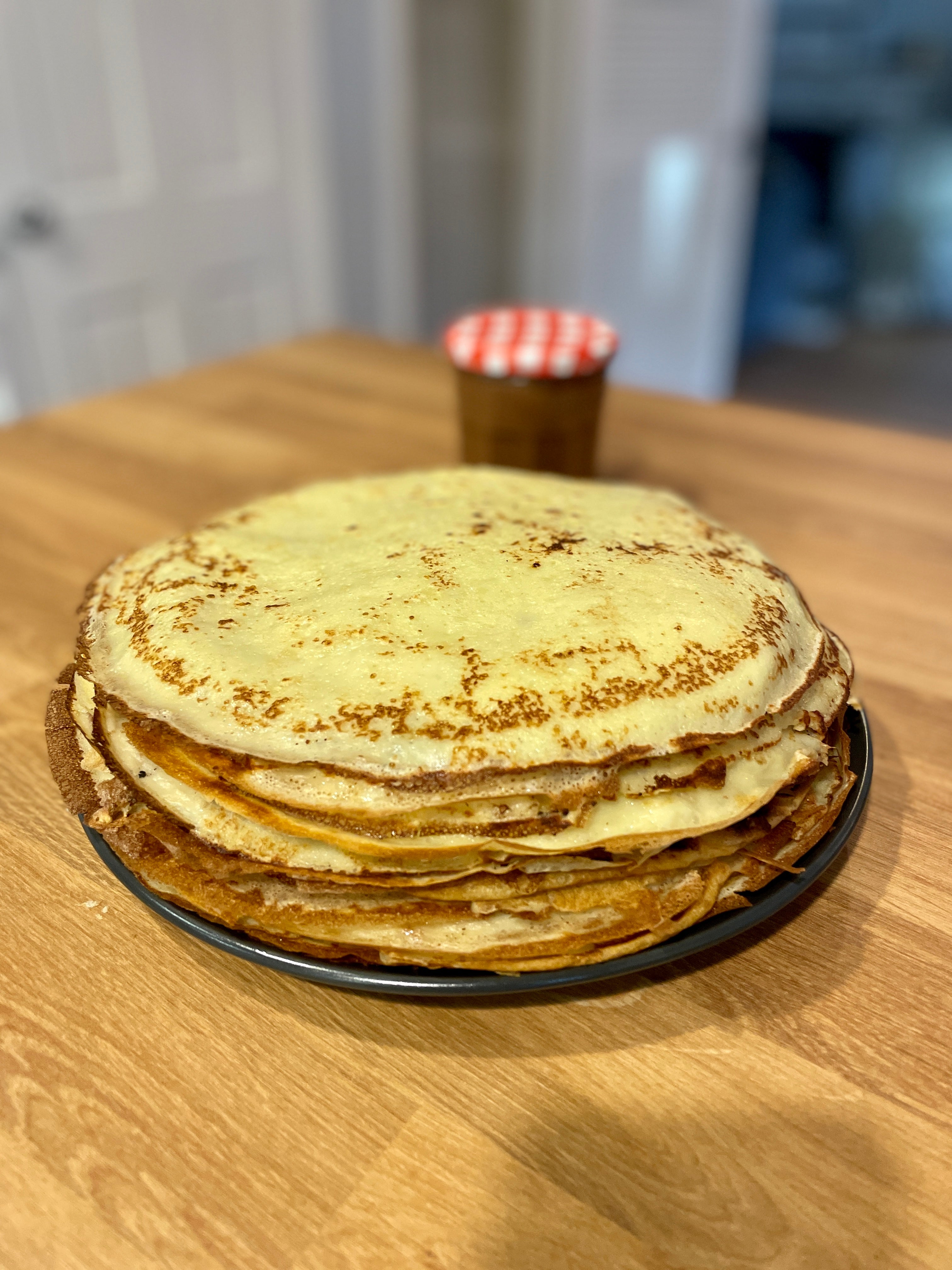A stack of freshly cooked French crêpes on a black plate, set on a wooden table with a jar of jam in the background.